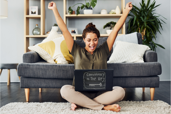 A young woman sat on the floor of her living room with her back against the sofa. She's sitting cross legged and has a laptop perched on her lap. On the laptop there is a sign which says consistency is key. The image has been used to portray how to create a simple blogging routine you'll actually stick to.