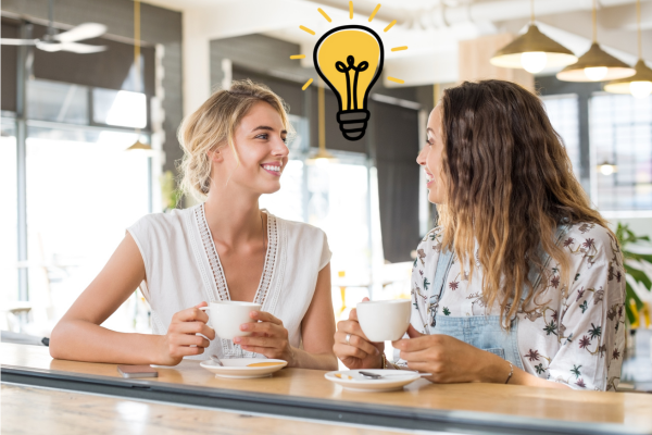 two women chatting at a table with cups of coffee. It's a casual conversation but the lightbulb between their heads show how to find blog post ideas in everyday conversations